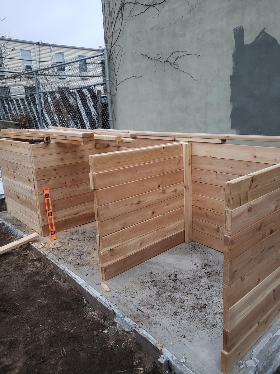 Cedar plank walls being framed into a two-bay composting bin on the concrete pad